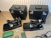 Front view of two Singer Featherweight sewing machines on bases with two black hard shell carrying cases behind
