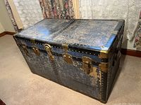 Full view of the large rectangular vintage baggage trunk with worn blue decorative finish, intact leather straps, metal corner reinforcements, and brass hardware.