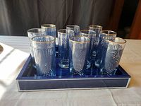 Set of 10 high ball glasses arranged inside matching deep blue glass tray showing gold dipped rims and deep blue decorative swirls on glasses.