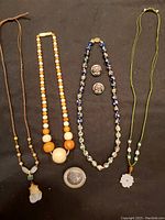 Photo showing six individual jewelry pieces laid out on black background: two carved Chinese jade necklaces, one cloisonné bead necklace, matching clip-back earrings, a strand of graduated orange and cream beads, and one sterling silver stamped warrior brooch/pendant.