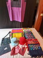 Photo showing the entire collection with the purple Huipil top hanging, scall scarves/table runners neatly folded, three drawstring gift bags, and two baby hats placed on a table.