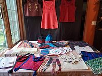 Overview of various handwoven and embroidered Mayan Indian textile items laid on a table and hanging against a dark background, including clothing, bags, scarves, and small accessories.