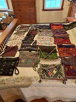 Photo showing a collection of 19 handwoven woolen tote bags arranged on a large bed with varied traditional Mayan patterns and colors including purples, reds, browns, greens, and oranges.
