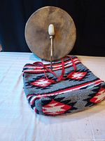 Full view of the elk skin drum with drumstick standing behind a Southwest patterned wool tote placed on a table.