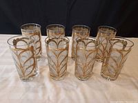 Set of eight Georges Briard highball glasses arranged in two rows, showing gold decorative pattern and clear glass body on white cloth background.