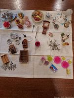 Full view of assorted miniature dollhouse accessories laid out on cloth including food items, kitchenware, clocks, and decorative pieces.