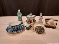 Seven vintage items displayed including porcelain teacups, figurines, a cork sculpture in glass, and a cloisonné trinket box.