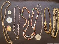 All nine pieces of jewelry displayed on a black background: two heavy gold-colored chains, multiple necklaces including the long multi-colored agate Beggar's necklace, the tri-strand tiger's eye bead necklace with gold discs, polymer clay bead necklace, pewter Scandinavian necklace with large circular etched pendant, jade disc pendant earrings, and a colorful enamel 'House' pin.
