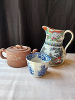 Full view of the three items showing the red clay tea pot with chipmunk lid, colorful pottery pitcher, and blue and white tea cup.