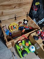 Open wooden toy box showing contents including Woody doll, Mickey Mouse plush, cars, and other assorted toys.