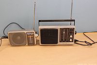Two vintage portable AM/FM radios side by side, showing front and side views with telescopic antennas extended and power cords visible.