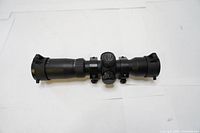 Full horizontal view of black Excalibur Tact-100 rifle scope on white background showing eyepiece, turret and objective lens caps closed.