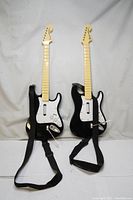 Pair of black Fender Stratocaster style guitar controllers with attached straps against a white backdrop