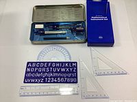 Photo of full set laid out with blue metal box, compass, protractor, alphabet stencil, 2 triangle rulers, ruler, eraser and sharpener.
