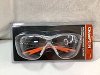 Front view of the ballistics shooting glasses in original packaging showing clear lens and orange arms with Champion branding on header card.