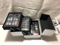 Top down view of open travel makeup case showing trays containing eyeshadow palettes, lipsticks, mascara, nail polishes, and cosmetic jars