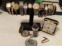 Full front view of 12 vintage wristwatches on display, two compasses, travel alarm clock, and two small pocket cutting utensils on table.