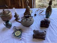 Wide view of all items in the lot arranged on a table including two lamps, kettle, carved cat figurine, blue goblet, oil lamp, and decorative metal pieces.