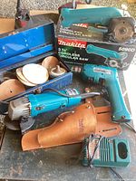 Makita angle grinder, cordless driver, sanding discs, charger, and tool box on a table next to the cordless circular saw and its box.