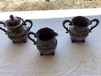 Photo of three silver-plated tea set pieces: lidded sugar canister, creamer/milk pitcher, and urn with double handles, all tarnished and on a white cloth