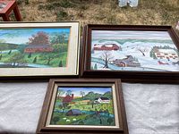 Set of three framed paintings showing spring, winter, and summer rural farmhouse scenes on a white quilt background outdoors.