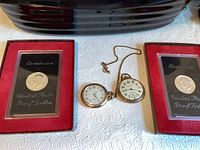 Two gold plated pocket watches and two Eisenhower proof silver dollars displayed on white textured surface