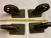 Four vintage heavy cast iron wheels with mounting plates arranged on a white surface, showing the side view with a measuring tape indicating approximate dimensions.