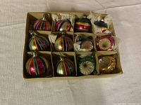 Full view of the 12 vintage glass Christmas ornaments in divided cardboard box, showing spherical and bulb shapes with striped colorful patterns and reflective centers.