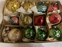Overall view of 12 vintage glass Christmas ornaments in cardboard divider box, showing variety of colors and designs wrapped in tissue paper.