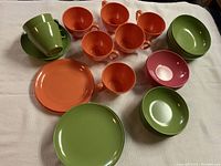 Top view of colorful orange, green, and pink Mel Mac melamine cups, bowls, and plates arranged on a cloth surface.