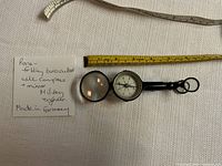 Photo of binoculars folded with compass and mirror visible, showing a yellow measuring tape for scale and a note indicating it is a rare folding binocular set made in Germany for military engineers.