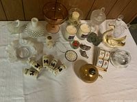 Overview of lot showing various clear and amber glassware, candles, and figurines arranged on a table covered with white cloth.