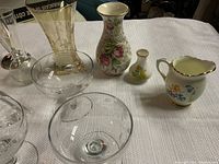 Photo showing mix of clear glass stemmed bowls, decorative glass items, two floral ceramic vases, and two small floral creamers on a white textured cloth.