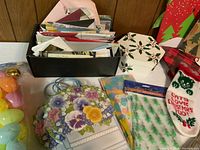 Photo showing multiple greeting cards stored in a black box, several large floral gift bags with pastel flowers and light blue handles, a small Christmas stocking for cats, and various holiday gift bags arranged around the box