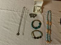 Photo showing multiple jewelry items including necklaces, bracelets, a brooch, and a jewelry box on a light-colored fabric surface.