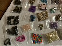 Various beads in small ziplock plastic bags arranged on a cloth surface showing metal, shell, plastic, and glass beads