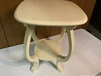 Front view of the small cream-colored wooden side table showing its rounded rectangular top, curved legs and the round lower shelf.