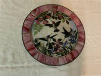 Photo of round stained glass panel showing painted scene of hummingbirds and flowers, with pink and purple stained glass border.