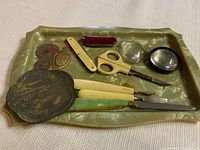 Photo of green metal tray with assortment of vintage tools and accessories including scissors, knives, magnifying glasses, and decorative items.