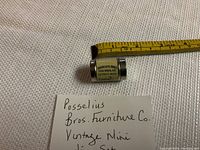 Small cylindrical tube container with label and black cap, placed alongside a measuring tape to show compact size. Handwritten note in view describes item as Posselius Bros. Furniture Co. vintage mini dice set.