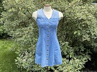 Front view of sleeveless light blue denim dress with button-up closure and chest pockets, displayed on mannequin outdoors against green foliage.