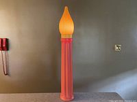 Full view of a tall red blow mold candlelight decoration with an orange flame top obstructing a wall with a light switch.