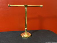 Front view of polished brass hand towel rack stand with octagonal base and horizontal bar against red background.