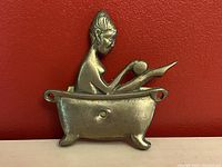 Front view of small brass wall decoration showing a lady sitting in clawfoot bathtub, with visible mounting holes.