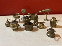 Collection of miniature brass household items, including a telescope, BBQ grill, and assorted kitchen pots and pans, arranged on a red background.