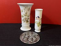 Two floral vases, one larger with beige flowers and one smaller with bird motif, alongside a round glass flower frog.