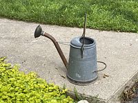 Side view of galvanized watering can placed on cement near some plants showing overall shape, metal surface, spout and handles.