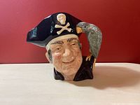 Front view of the Long John Silver Royal Doulton Toby jug showing detailed face and black pirate hat with skull and crossbones, and grey bird handle attached to shoulder.