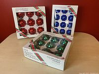 Three unopened boxes of vintage Noma Double Glow faceted glass Christmas ball ornaments in red, blue, and green.