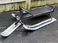 Side view of NOMA Super GT Racer Toboggan Sled showing black seat, metal frame, and metal skis with some rust.